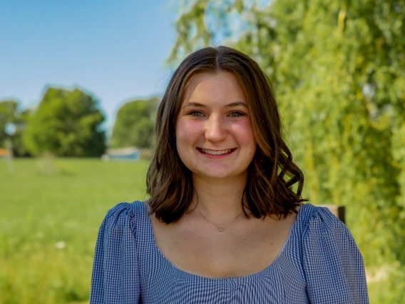 A person with brown hair smiles outdoors, wearing a checkered top and blue jeans, with a green field and trees in the background.