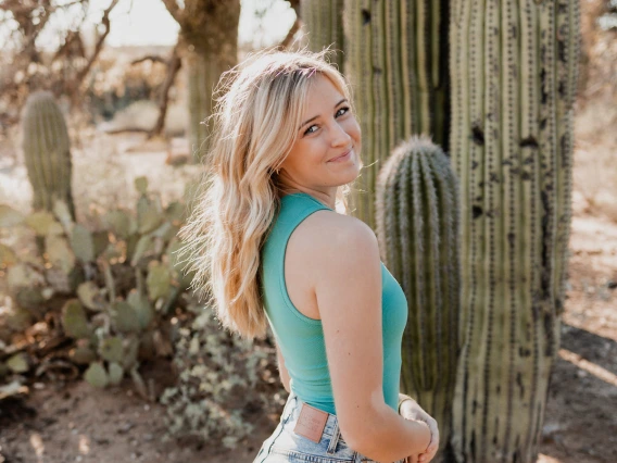 A person standing by a cactus in a sunlit desert landscape, wearing a turquoise top and blue jeans.