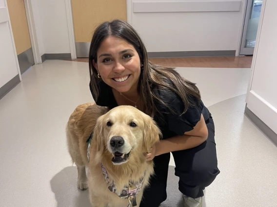 A person with long brown hair in black scrubs kneels beside a golden retriever in a hallway.
