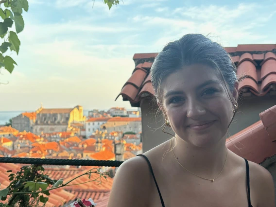 Person standing over a balcony with several buildings behind them and leaves from a green tree