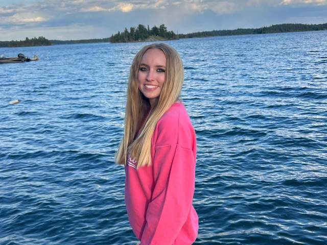 Person with long blonde hair in a pink sweatshirt and jeans, smiling in front of lake scene.