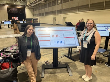 Two people stand beside a large digital poster display in a convention hall. The poster features charts and text