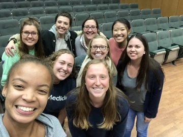 A group of eight people smiling and taking a selfie in an empty lecture hall.