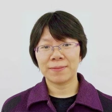 A person with short dark hair and purple glasses, wearing a purple jacket.