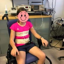 A child is sitting in a chair indoors, wearing respiratory bands around her chest and abdomen.