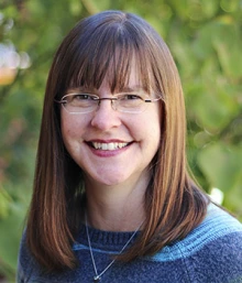 A person with long brown hair and glasses, smiling, wearing a blue sweater. The person is in front of a leafy green background.