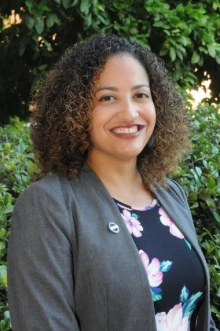 A person with curly hair smiles, wearing a gray blazer and a floral dress, with green foliage in the background.